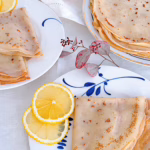 Апетитні французькі тонкі млинці, складені трикутничками на керамічній тарілці, щедро посипані цукром, политі свіжим лимонним соком та прикрашені зеленими листочками м'яти. Поруч лежать половинки свіжого лимона.