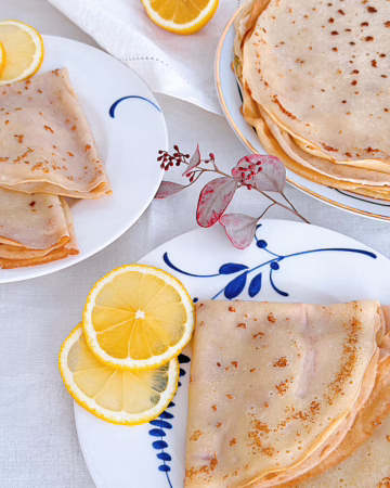 Апетитні французькі тонкі млинці, складені трикутничками на керамічній тарілці, щедро посипані цукром, политі свіжим лимонним соком та прикрашені зеленими листочками м'яти. Поруч лежать половинки свіжого лимона.