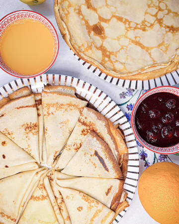 Тонкі золотисті данські млинці pandekager, складені стопкою на красивій тарілці, поруч стоїть баночка з ягідним варенням та чашка гарячої кави