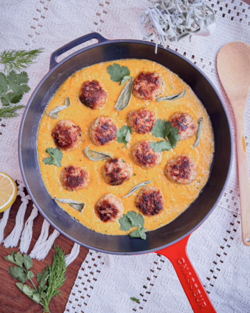 Апетитні золотисто-коричневі фрикадельки з індички, що тушкуються у яскраво-помаранчевому кремовому гарбузово-шавлієвому соусі у великій круглій чавунній сковорідці червоного кольору. Зверху страва прикрашена свіжим листям кінзи та сріблястою шавлією, поруч на білій мереживній скатертині лежать дерев'яна лопатка, половинка лимона та гілочка кропу.
