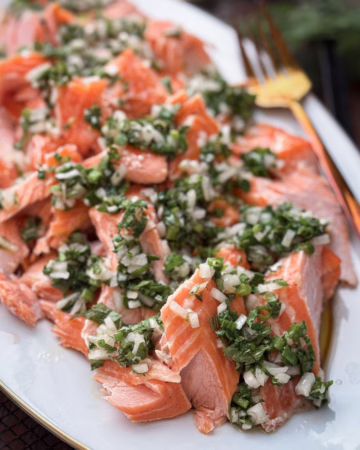 Великі шматки томленого лосося, щедро политі зеленим трав'яним соусом з оливкової олії та шалоту, подані на світлій керамічній тарілці ручної роботи.