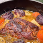 Цілий шматок томленого свинячого ошийка в густому соусі з великими шматками запеченої картоплі та моркви, поданий у керамічній тарілці та прикрашений гілочками свіжого чебрецю.