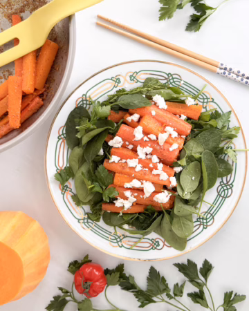 Тарілка з яскравими карамелізованими овочами: смажений гарбуз, батат та морква в апельсиновій глазурі, подані на свіжому шпинаті з шматочками сиру фета.