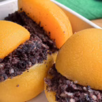 Половинки жовтих абрикосів, між якими викладена темна начинка з розім'ятої кров'яної ковбаси. Фаршировані фрукти лежать у білій керамічній формі для запікання на фоні зеленої лляної серветки.