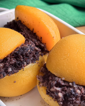 Половинки жовтих абрикосів, між якими викладена темна начинка з розім'ятої кров'яної ковбаси. Фаршировані фрукти лежать у білій керамічній формі для запікання на фоні зеленої лляної серветки.