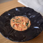 Чорна керамічна тарілка з рельєфними краями наповнена густим вершковим рагу з білої квасолі, томатів черрі та свіжого шпинату на дерев'яному столі поруч із лляною серветкою.