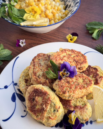 Естетична подача золотистих курячих кнелей на вінтажній білій тарілці з синім розписом виробництва Коросненського порцелянового заводу. Страва прикрашена квітами фіалок, свіжою м'ятою та скибочкою лимона; на фоні — піала з кукурудзою та м'ятою.