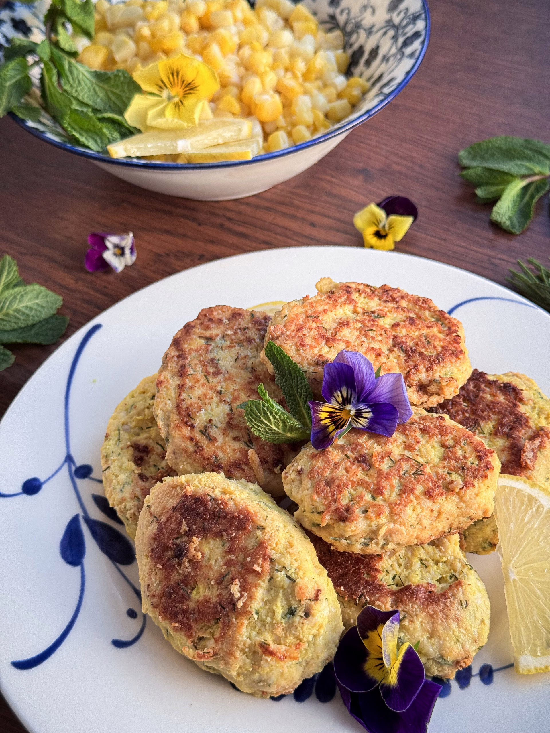 Естетична подача золотистих курячих кнелей на вінтажній білій тарілці з синім розписом виробництва Коросненського порцелянового заводу. Страва прикрашена квітами фіалок, свіжою м'ятою та скибочкою лимона; на фоні — піала з кукурудзою та м'ятою.