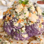 Ціла запечена золотиста цвітна капуста, щедро полита вершковим соусом з молюсками, каперсами та зеленню, на елегантній світлій керамічній тарілці.