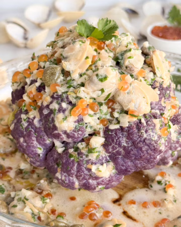 Ціла запечена золотиста цвітна капуста, щедро полита вершковим соусом з молюсками, каперсами та зеленню, на елегантній світлій керамічній тарілці.