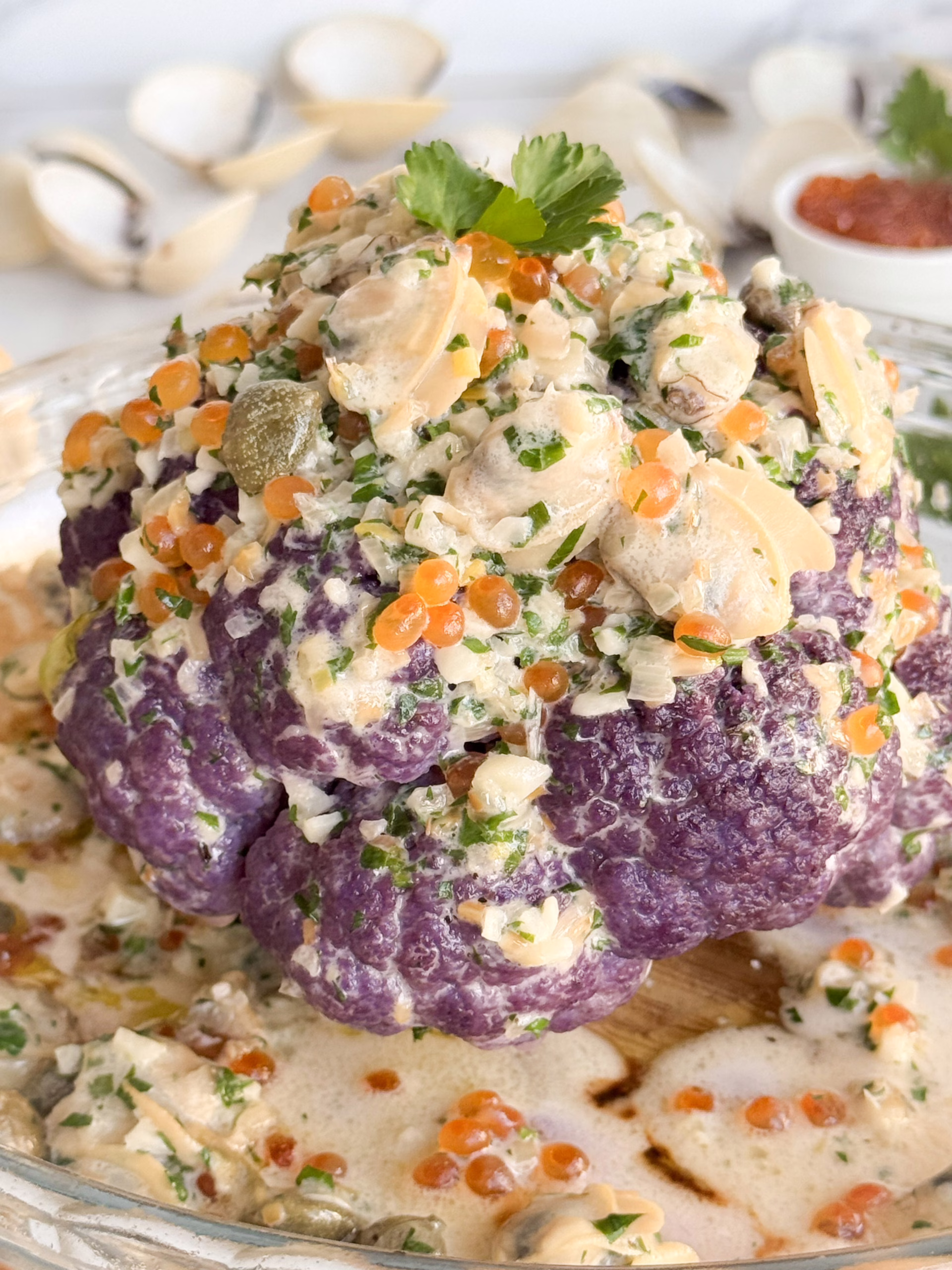 Ціла запечена золотиста цвітна капуста, щедро полита вершковим соусом з молюсками, каперсами та зеленню, на елегантній світлій керамічній тарілці.