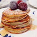 Висока стопка рум'яних бананових оладок на світлій вінтажній тарілці, щедро полита кленовим сиропом і прикрашена ягодами та кокосовою стружкою.