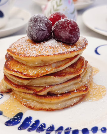 Висока стопка рум'яних бананових оладок на світлій вінтажній тарілці, щедро полита кленовим сиропом і прикрашена ягодами та кокосовою стружкою.