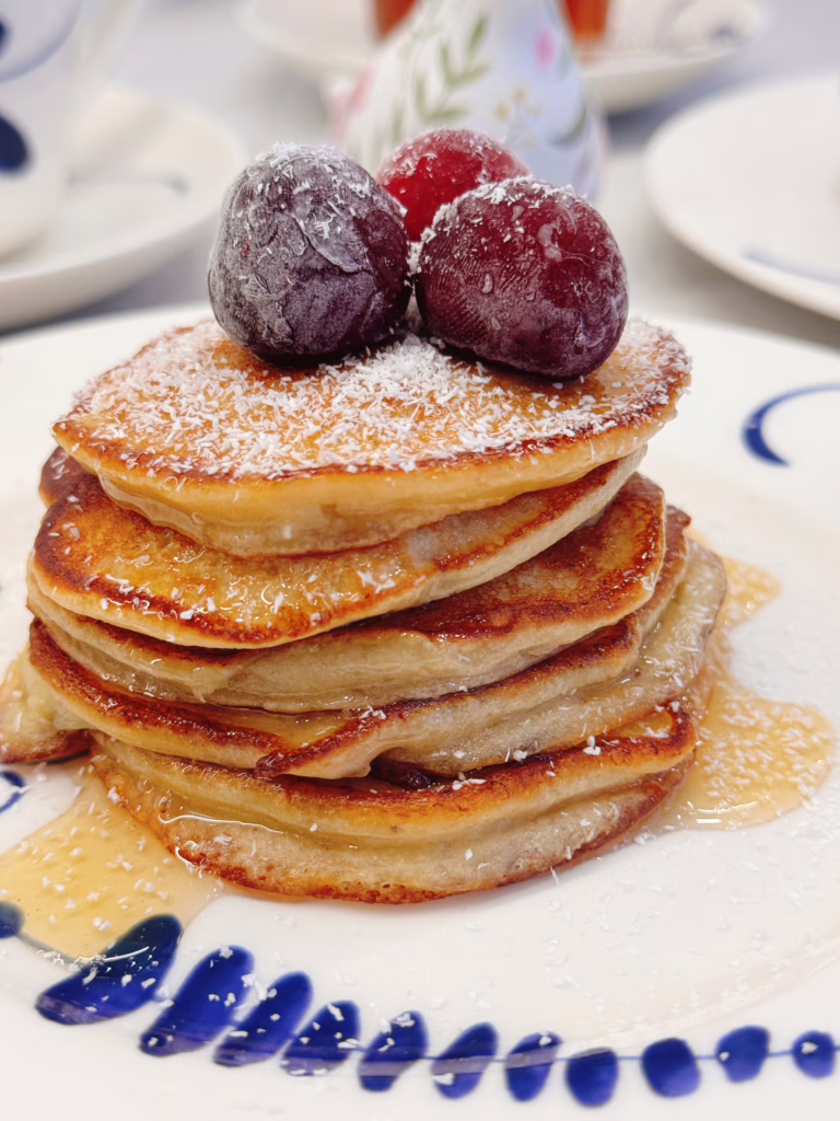 Висока стопка рум'яних бананових оладок на світлій вінтажній тарілці, щедро полита кленовим сиропом і прикрашена ягодами та кокосовою стружкою.