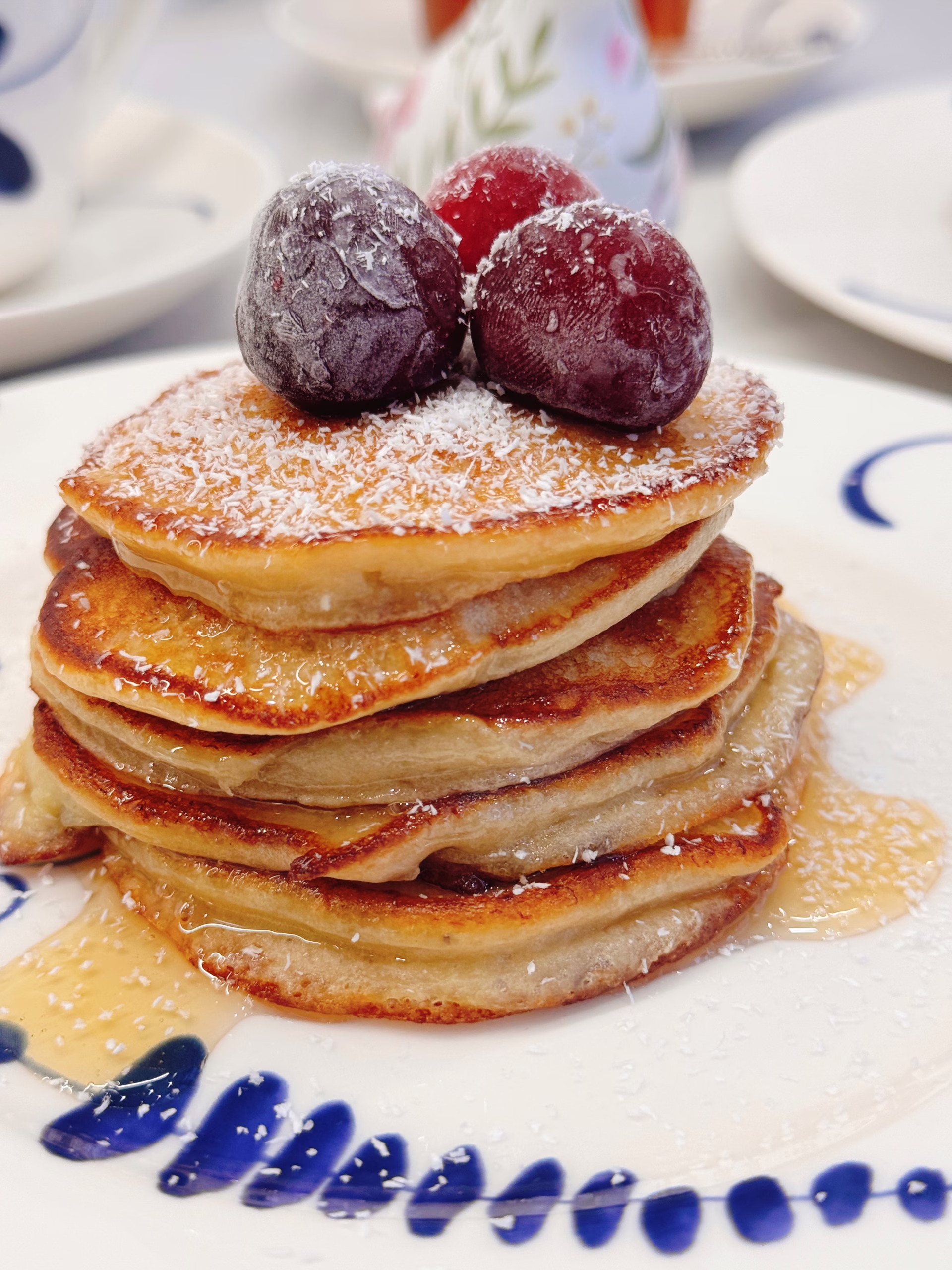 Висока стопка рум'яних бананових оладок на світлій вінтажній тарілці, щедро полита кленовим сиропом і прикрашена ягодами та кокосовою стружкою.
