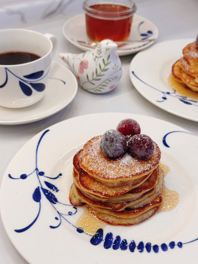 Висока стопка рум'яних бананових оладок на світлій вінтажній тарілці, щедро полита кленовим сиропом і прикрашена ягодами та кокосовою стружкою.