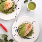 Шматочки золотистого курячого філе на подушці з зеленого горошку та бобів едамаме, политі яскравим смарагдовим м'ятним соусом і посипані сиром фета, подані на світлій керамічній тарілці.