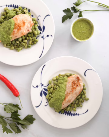 Шматочки золотистого курячого філе на подушці з зеленого горошку та бобів едамаме, политі яскравим смарагдовим м'ятним соусом і посипані сиром фета, подані на світлій керамічній тарілці.