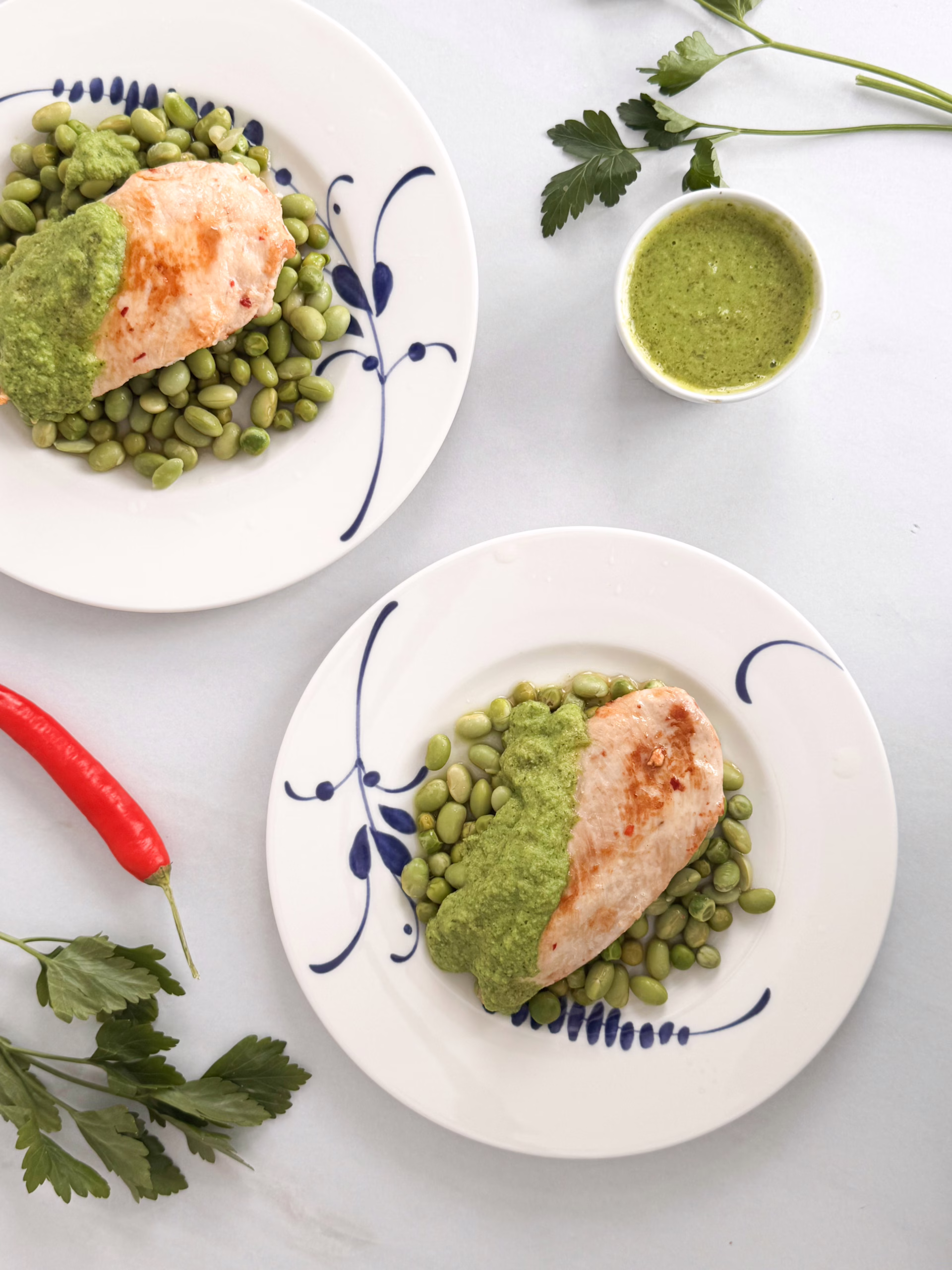 Шматочки золотистого курячого філе на подушці з зеленого горошку та бобів едамаме, политі яскравим смарагдовим м'ятним соусом і посипані сиром фета, подані на світлій керамічній тарілці.