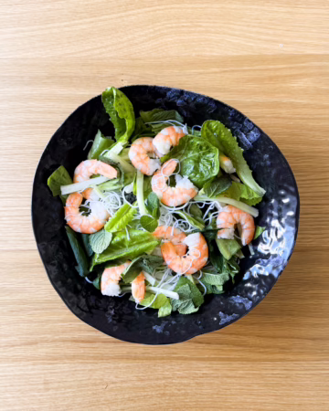 Глибока чорна керамічна тарілка з в'єтнамським боулом: рисова локшина, ромен, свіжа м'ята та рожеві креветки на дерев'яному столі.