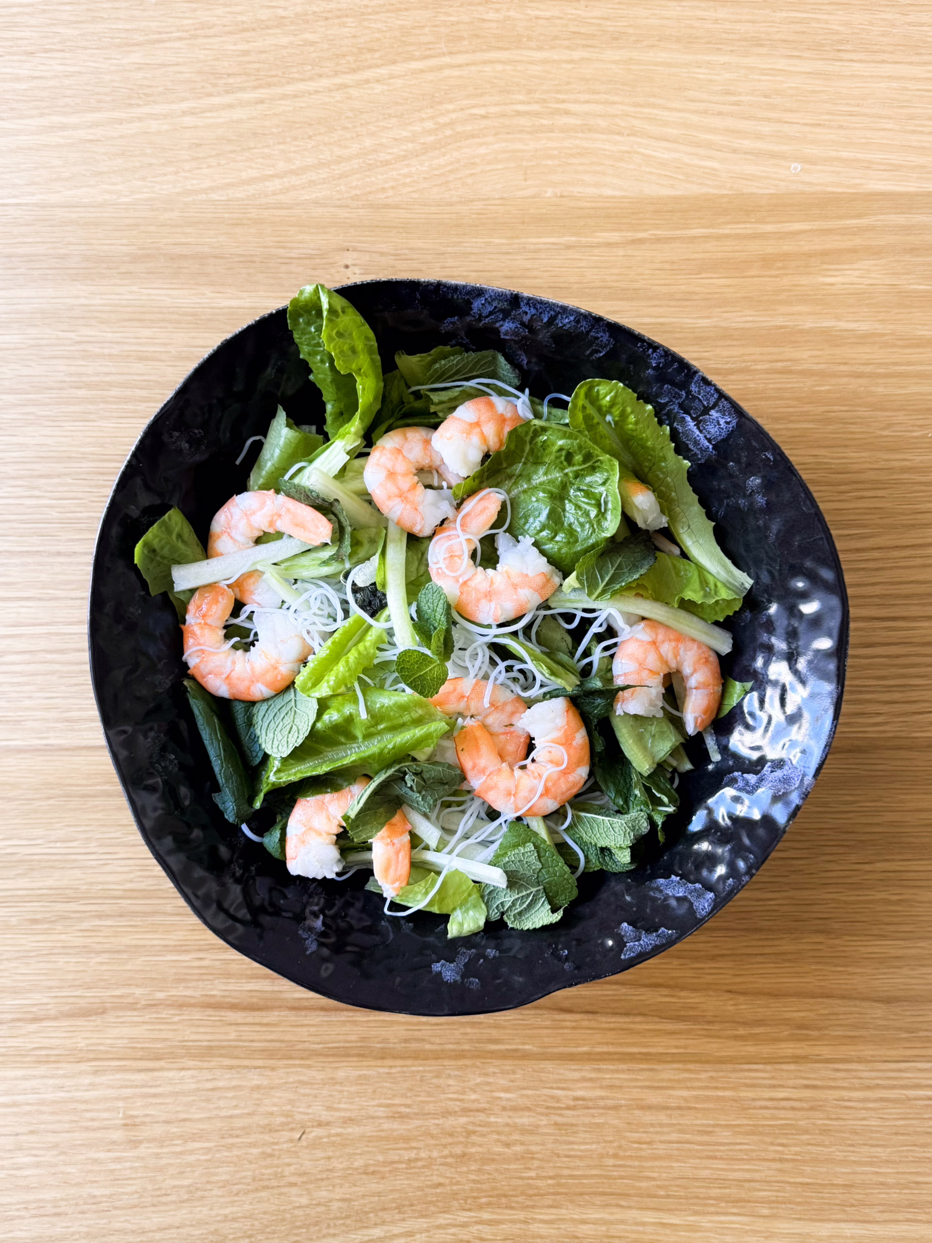 Глибока чорна керамічна тарілка з в'єтнамським боулом: рисова локшина, ромен, свіжа м'ята та рожеві креветки на дерев'яному столі.