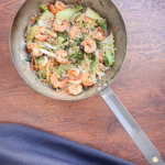 Крупний план металевої миски (вока), наповненої гарячим стір-фраєм із великими рожевими креветками, зеленими овочами (пак-чой, броколіні, селера), помаранчевою морквою та щедро посипаним подрібненим арахісом. Миска стоїть на дерев'яному столі.