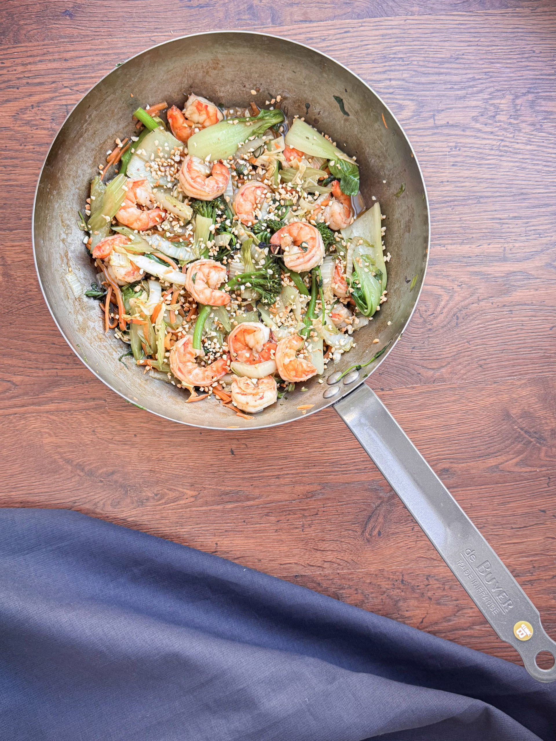 Крупний план металевої миски (вока), наповненої гарячим стір-фраєм із великими рожевими креветками, зеленими овочами (пак-чой, броколіні, селера), помаранчевою морквою та щедро посипаним подрібненим арахісом. Миска стоїть на дерев'яному столі.