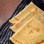 Тонкі золотисті данські млинці pandekager, складені стопкою на красивій тарілці, поруч стоїть баночка з ягідним варенням та чашка гарячої кави