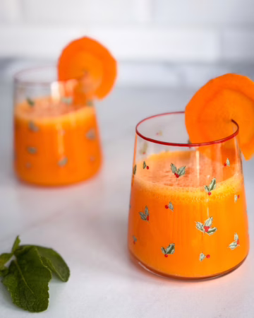 морквяний лимонад з медом та кубиками льоду у високій склянці, прикрашений скибочкою свіжої моркви та гілочкою м'яти.
