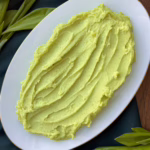 Гладка хвиля зеленого картопляного пюре на світлій керамічній тарілці з тонким золотим кантом; свіже листя черемші та шалот на дерев'яній дошці; процес вмішування зелених вершків у протерту картоплю.