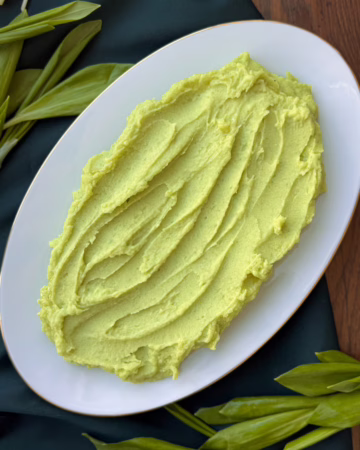 Гладка хвиля зеленого картопляного пюре на світлій керамічній тарілці з тонким золотим кантом; свіже листя черемші та шалот на дерев'яній дошці; процес вмішування зелених вершків у протерту картоплю.