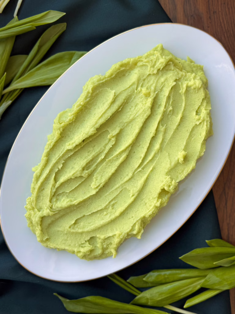 Гладка хвиля зеленого картопляного пюре на світлій керамічній тарілці з тонким золотим кантом; свіже листя черемші та шалот на дерев'яній дошці; процес вмішування зелених вершків у протерту картоплю.
