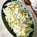 Хрусткий весняний салат з нарізаною півкільцями редискою та молодою капустою на темно-зеленій тарілці із золотою облямівкою.