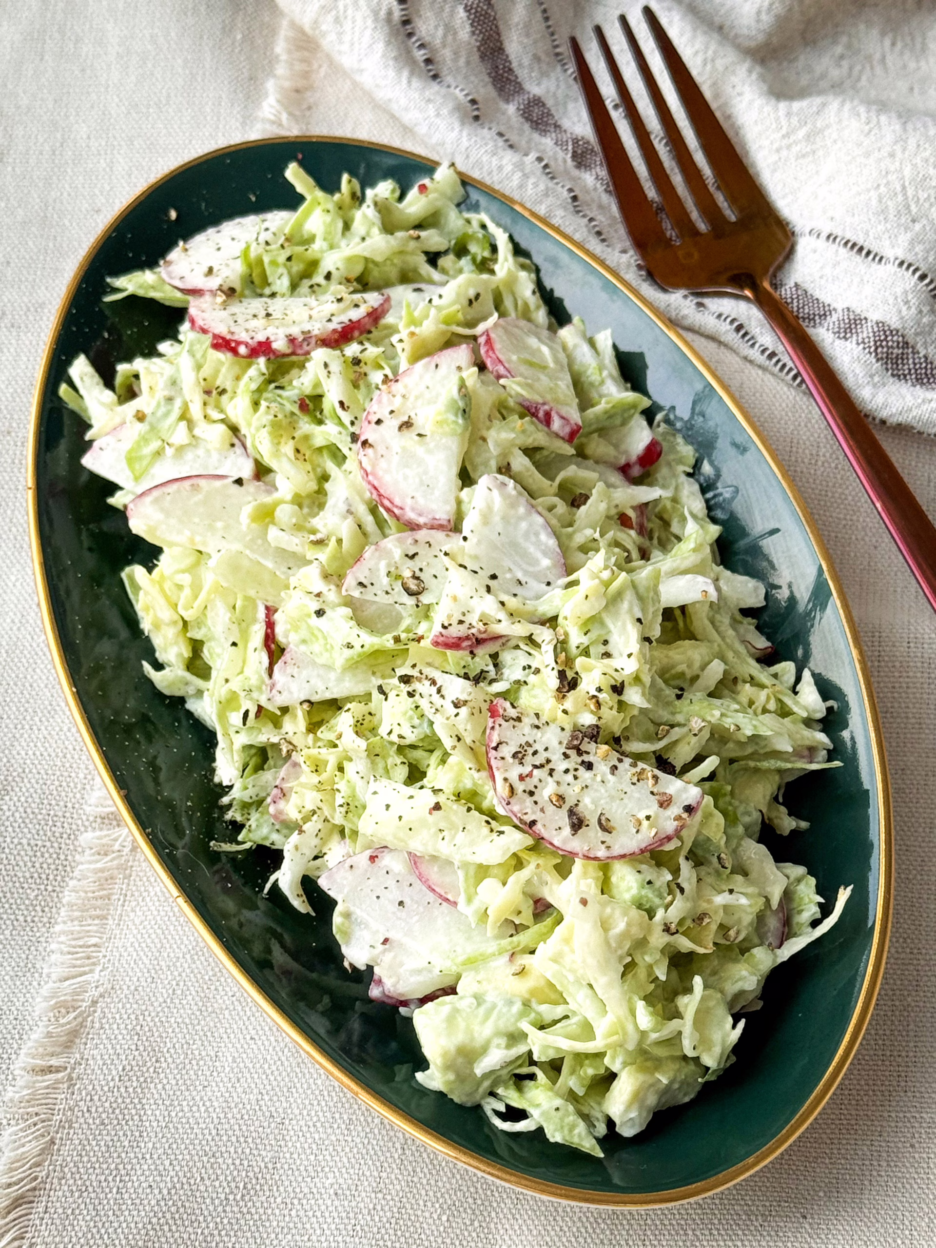 Хрусткий весняний салат з нарізаною півкільцями редискою та молодою капустою на темно-зеленій тарілці із золотою облямівкою.