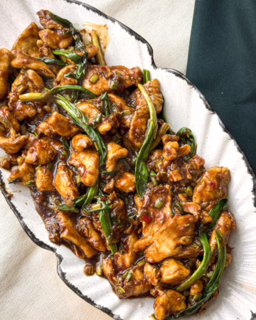 Тонкі скибочки курячого філе в густому глянцевому соусі поруч зі смарагдовою черемшею; процес додавання соєвої глазурі до обсмаженої курки на пательні.