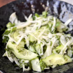 Тонко нарізаний білий фенхель та зелені півкільця огірка з кропом і петрушкою у темній фактурній керамічній мисці.