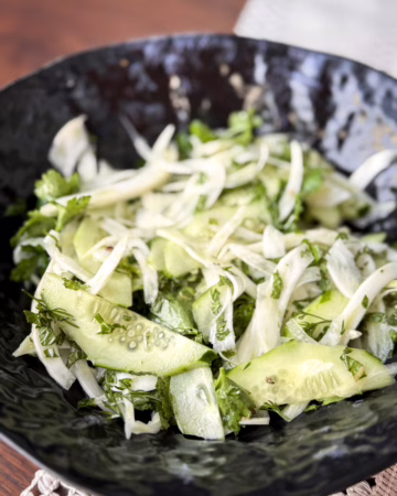 Тонко нарізаний білий фенхель та зелені півкільця огірка з кропом і петрушкою у темній фактурній керамічній мисці.