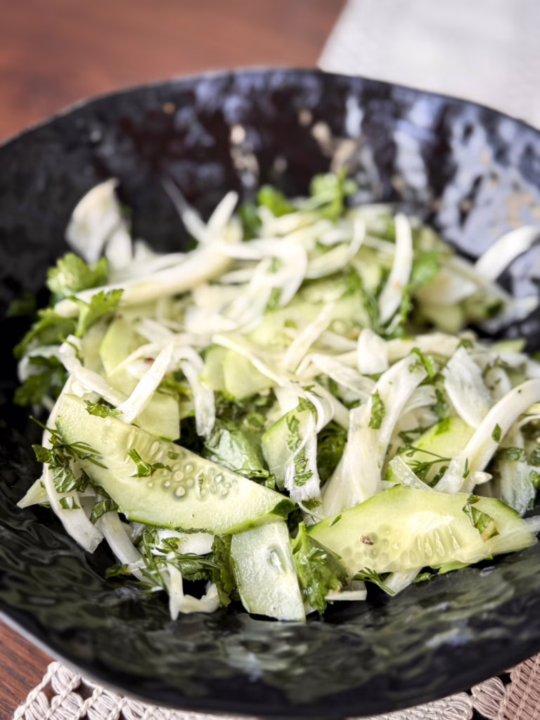 Тонко нарізаний білий фенхель та зелені півкільця огірка з кропом і петрушкою у темній фактурній керамічній мисці.