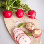 Естетичний flat lay сніданку: дерев'яна дошка зі скибочками багета, маленькою керамічною мисочкою з вершковим маслом кімнатної температури, свіжим червоним редисом з невеликими зеленими хвостиками та пластівцями морської солі. Природне світло.