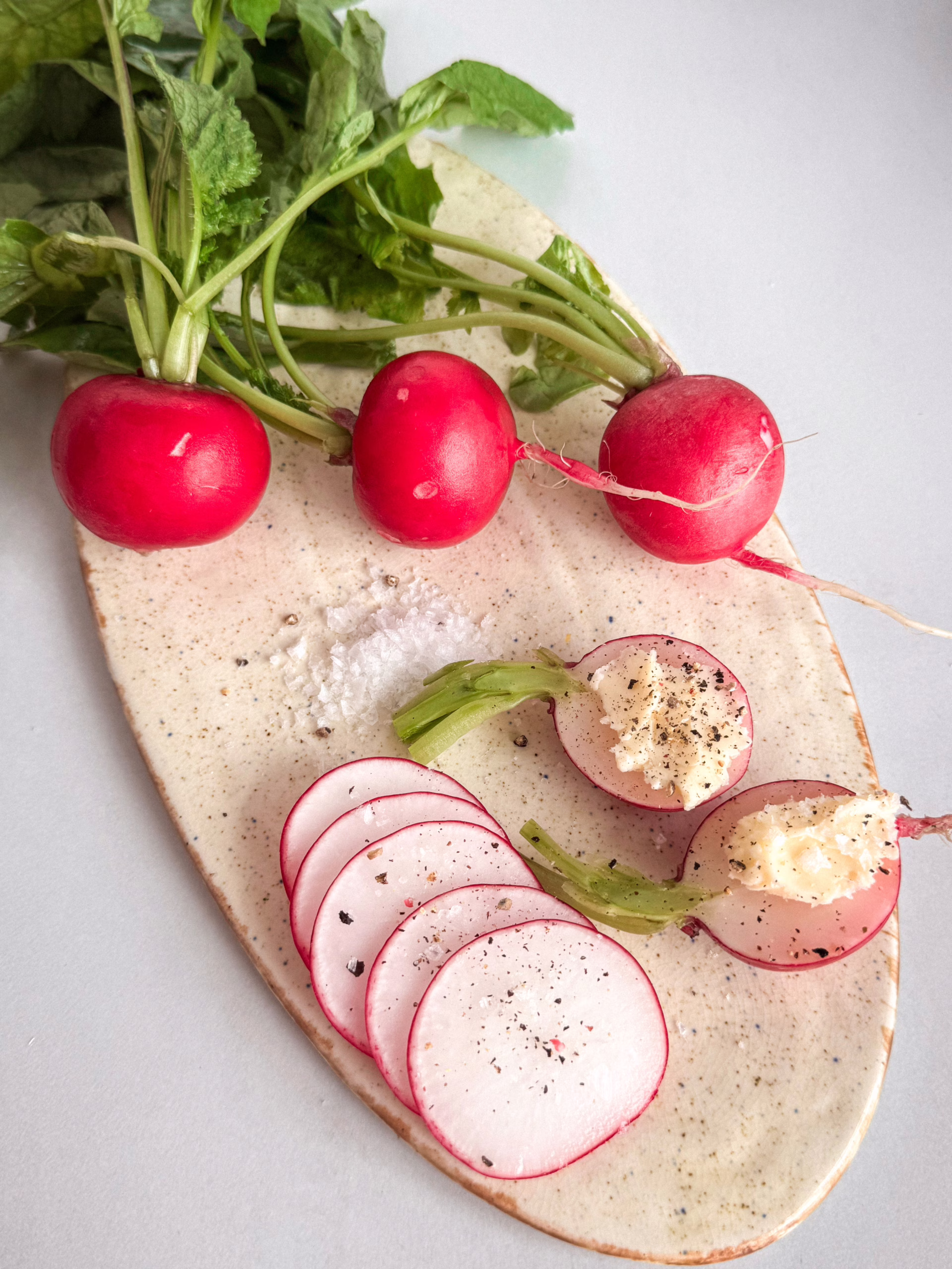 Естетичний flat lay сніданку: дерев'яна дошка зі скибочками багета, маленькою керамічною мисочкою з вершковим маслом кімнатної температури, свіжим червоним редисом з невеликими зеленими хвостиками та пластівцями морської солі. Природне світло.