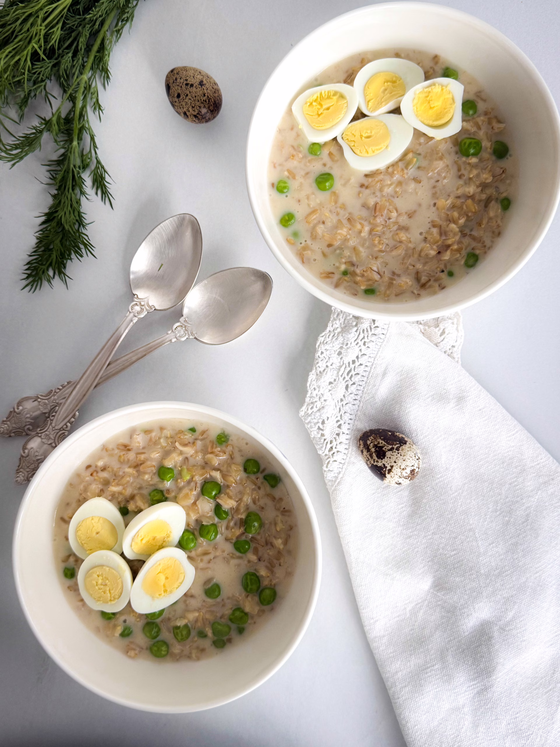 Дві білі керамічні піали з солоною кремовою вівсянкою, зеленим горошком та половинками перепелиних яєць, поруч срібні ложки та біла лляна серветка.